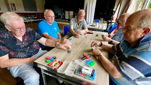 A group of five men play poker around a table.