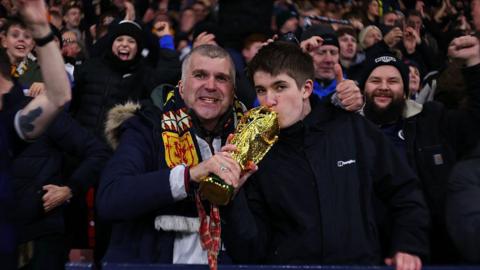  A crowded football stadium with fans celebrating. Two people in the foreground hold a golden trophy and wear scarves with a red lion emblem.