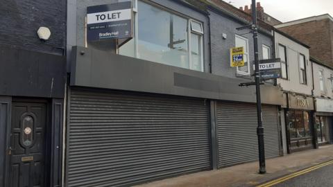 The site which has been given approval. A disused commercial front with black shutters, with an upper story with windows facing outwards. Next door are other businesses on the street. "To let" signs are posted outside the space.