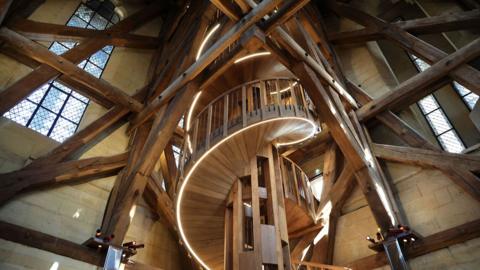 A spiral oak staircase stretching up through old style wooden beams.