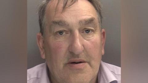 A mugshot of a man with dark grey hair. He wears a light pink shirt.