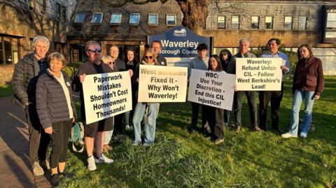 A group of people stand in front of a Waverley Borough Council sign on a grassy area with building in the background. They are holding signs which read "paperwork mistakes shouldn't cost homeowners thousands" and "WBC: Use your discretion - end this CIL injustice".