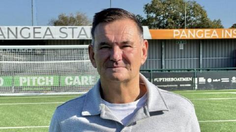 Paul Doswell standing on a football pitch, dressed in a light grey button-up shirt over a white top. Behind him, there is a goalpost and a building with a sign for Pangea Insurance. 
