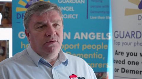 Mark Aldred with grey hair wearing a blue shirt with a Good Deeds Trust logo on it. He stands in front of a Guardian Angel advertising board. 