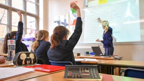 Two school girls wearing blazers put their right hands up in a class room while a teacher wearing a tracksuit points at something on a whiteboard.