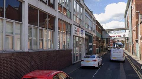 A narrow street, with a two-storey office building on the left side, and a three-storey redbrick building on the right. There are cars parked along the left side of the street.