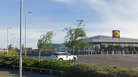The Lidl supermarket is in the background, and the car park which is almost empty in the foreground. The sun is low and the sky is blue with whispy cloud.