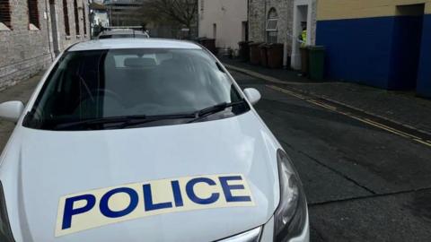 A police car parked in Rendle Street with a scene guard standing in the door way of a house behind police incident tape. 