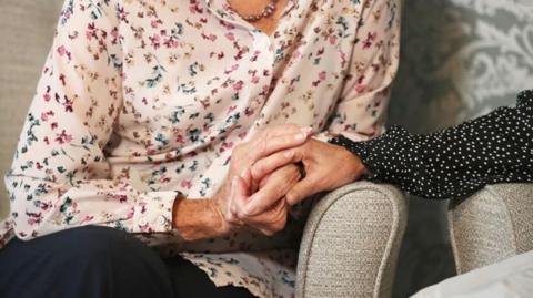 A hand clasped around the arm of an elderly woman. both are leaning on the arms of grey armchairs. The arm of the person on the right is clad in the sleeve of a black blouse with small white polka dots. The elderly lady is wearing a light coloured blouse with a a green, yellow and pink floral pattern and dark trousers. She is also wearing a beaded necklace.