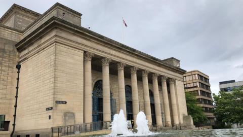 The exterior of Sheffield City Hall, a pale stone building with columns along the front entrance. A fountain is in operation directly outside the entrance.