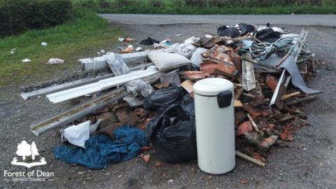 A pile of mixed waste including a pedal bin, masonry, guttering and pillows lies discarded off the side of a rural road.