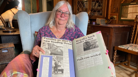 Penny is sitting in a chair with a patterned top on, long grey hair and a necklace while holding  an open scrap book of articles of flooding in Bewdley