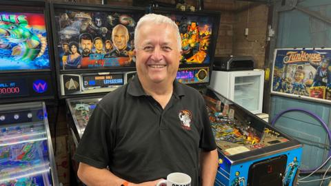 Mark Squires,  with short white hair, wearing a grey polo shirt, standing in front of three colourful pinball tables