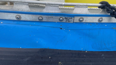 The image shows the side of a blue-painted boat hull with visible impact damage, including a small puncture hole and scuff marks along the metal panel. Above the damaged area, there is a metal railing and bolts with handwritten markings near one section, suggesting the damage has been inspected or noted.