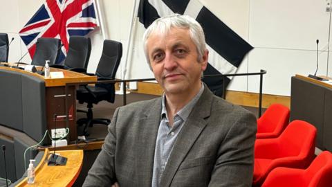 Dick Cole has grey grey hair and wears a grey jacket and black and white checked shirt. He stands inside the Cornwall Council chamber with seats and the Union and Cornish flags behind him. 