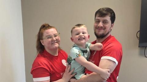 Libby and George stand in front of a white wall wearing their London Marathon T-shirts and holding son Ritchie between them. 