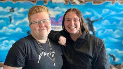 Two young women standing next to each other with one arm on the other's shoulder, smiling.