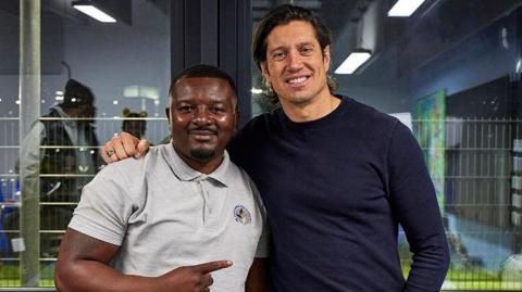 Be The Change founder Daniel Sukula smiles wearing a grey polo top and pointing his finger at a smiling 91ȱ broadcaster Vernon Kay who has his arm resting on the former's shoulder 