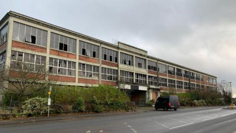 A dilapidated building on the outskirts of Dumfries with lots of broken windows and trees growing in front. A van is pulling in to turn at the main entrance.