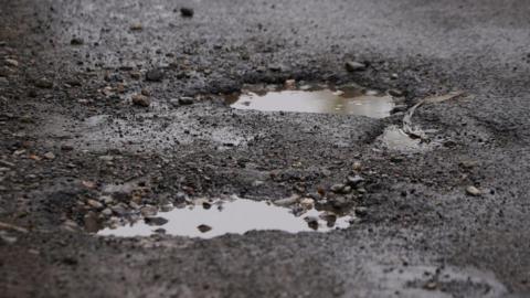 Two potholes in the road next to each other are filled with water. The road surface around them is breaking up.