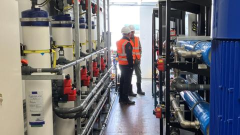 Two men in orange hi-vis jackets and white hard hats stand talking at the end of a corridor. Industrial pipework and cylinders line both walls of the passageway.