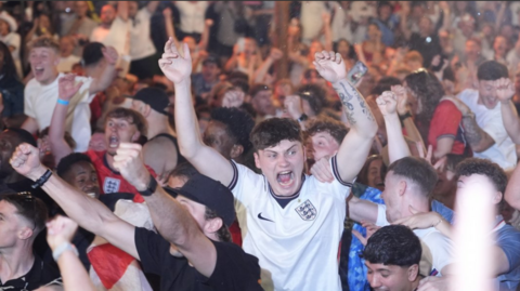 An England fan, in a white shirt, is held aloft by friends as he screams in delight
