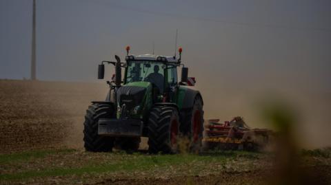 A green tractor in a dusty field, pulling equipment to plough the field. The sky in the distance is gloomy.