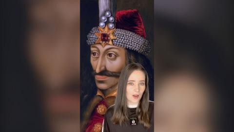 A woman in a black t-shirt with a mic clipped on to her is seen with a drawing of Vlad the Impaler behind her. He is wearing a red hat which has various gems on it.