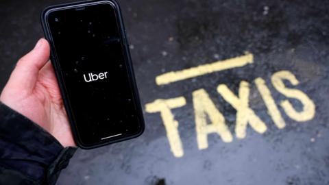 A person holding an apple i-phone with the uber app open, standing next to a yellow sign saying "taxi" on the road.