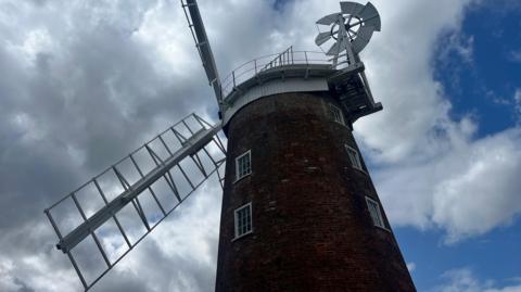 Dereham Windmill which stands tall with five floors. It has white sails and the photograph has been taken on a cloudy day with a blue sky.