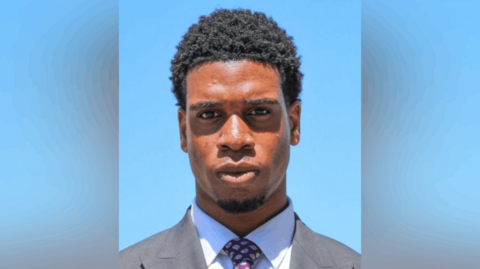 Divine Jacob, a man in a grey suit with a white shirt and purple tie against a blue background.