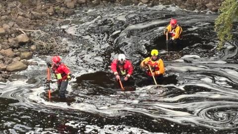 Rescue teams in hard hats and dry suits search the river Tyne in the Wylam area. The water is dark with foam swirling on the surface.
