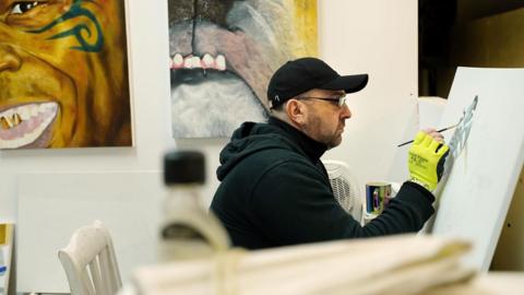 Mr Devonport sits at his easel, wearing a black cap, neon work gloves and hoodie. Two paintings can be seen adorning the wall next to him.