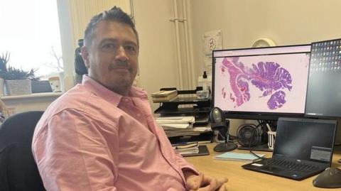 Consultant healthcare scientist James Woodland sits at a desk with computer screens seen behind him. He is wearing a pink shirt and looking at the camera.