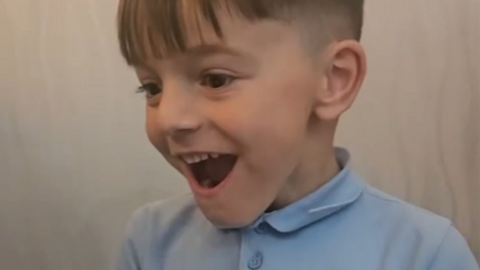 A six-year-old boy with light brown hair and wearing a light blue school polo-neck grins in delight.