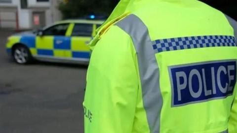 A police officer in hi-vis jacket taken from the back with a police car in the background