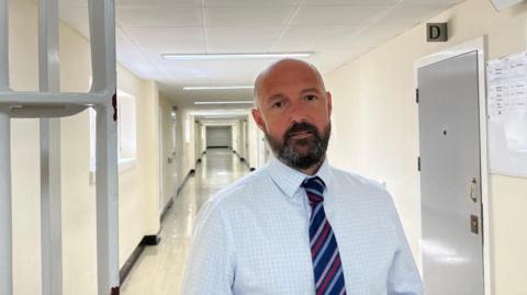 John De Carteret - A man with a bald head, and a black and grey beard looking at the camera. He's wearing a white and llight blue checked shirt, and a blue, pink, red and purple striped tie. He is standing next to some prison bars in a lonmg corridor of Guernsey's Les Nicolles prison.