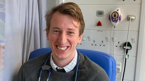 A doctor wearing a grey jumper with a stethoscope around his neck. He is sitting in a chair on a hospital ward