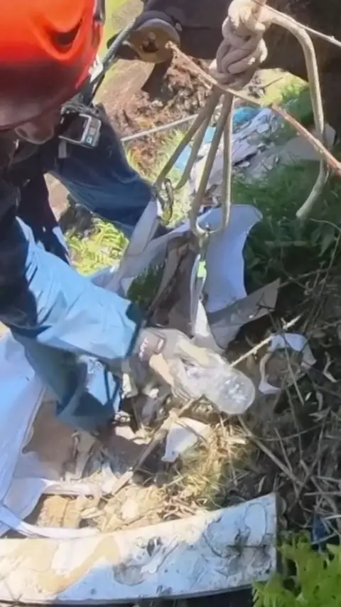 A man holding a rope putting a plastic bottle into a large plastic bag full of rubbish
