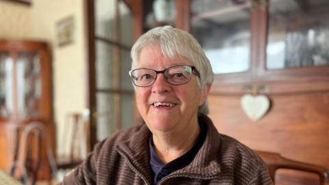 A white haired woman smiling towards the camera with black glasses and a brown jumper. 