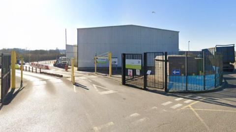 The image shows the outside of a recycling centre in Mole Valley. Some gates can be seen around the premises and a small blue sign with an arrow pointing inside.