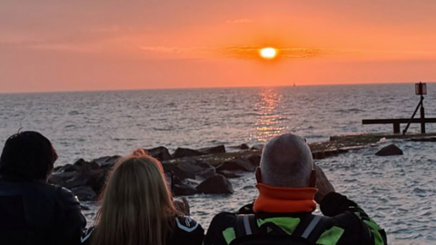 A group of three people viewed from behind look out at a vibrant orange and pink sunset over a calm sea, with rocks and a small wooden pier structure visible on the right.