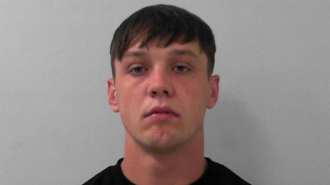 Patrick White's mugshot, wearing a black top and looking serious. He has short dark hair and stood in front of a grey background.