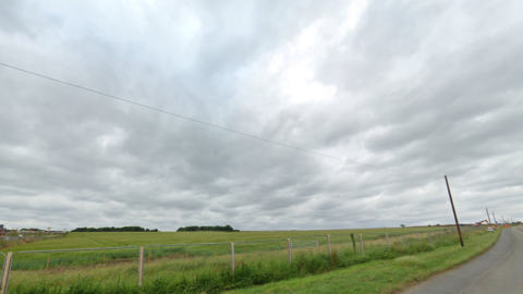 A green field to the side of a tarmac road. The field has long grass and a metal fence at the perimeter.