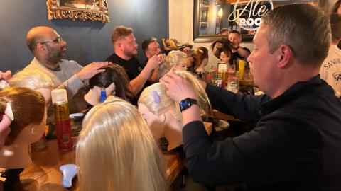 A group of men sitting around a long table. In front of them are dolls' heads attached to the tables, each with different colour hair. There is hairspray, brushes and clips on the table and the dads are playing with the hair.