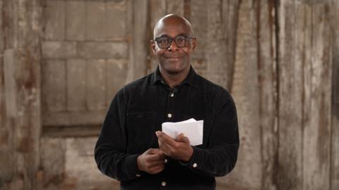 Paterson Joseph, a black man with a bald head, smiles at the camera. He is wearing black glasses and a black shirt. He is holding a piece of paper in his hand
