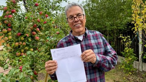 A man holds up a piece of paper as he stands in a garden. An apple tree with lots of fruit can be seen next to him. He wears a chequered shirt and glasses.