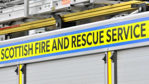 The words "Scottish Fire and Rescue Service" in black capital letters on a yellow background on the side of a fire engine. The engine is pictured in a close-up and only the grey shutters on its side and part of the ladders on its roof are visible.