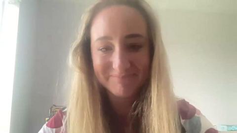 Screengrab from a zoom interview with a lady smiling into the camera, with long blonde hair, sat in front of a white wall. 