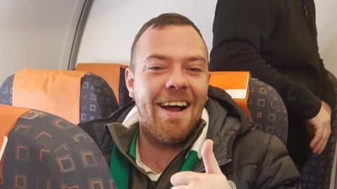 Kevin Davison, who is sitting in a plane seat, smiling at the camera and giving a thumbs up